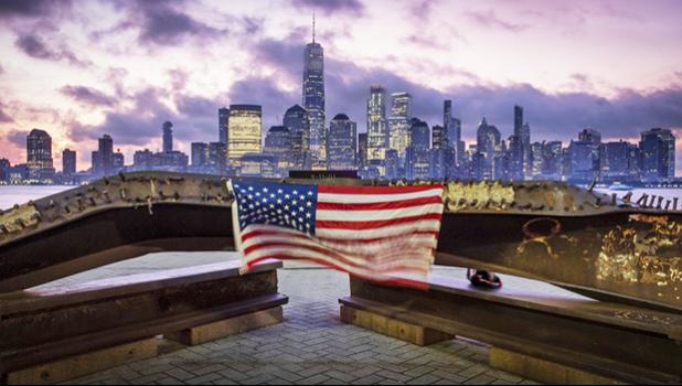 A U.S. flag hanging from a steel girder, damaged in the Sept. 11, 2001 attacks on the World Trade Center, blows in the breeze at a memorial in Jersey City, N.J., Sept. 11, 2019 as the sun rises behind One World Trade Center building and the re-developed area where the Twin Towers of World Trade Center once stood in New York City on the 18th anniversary of the attacks. (AP Photo/J. David Ake) A U.S. flag hanging from a steel girder, damaged in the Sept. 11, 2001